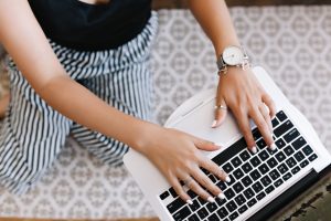 Woman in black and white trousers with white painted nails typing on a laptop keyboard.