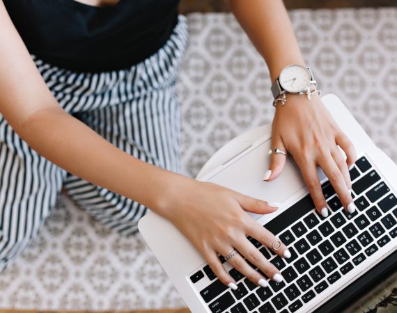 Woman in black and white trousers with white painted nails typing on a laptop keyboard.