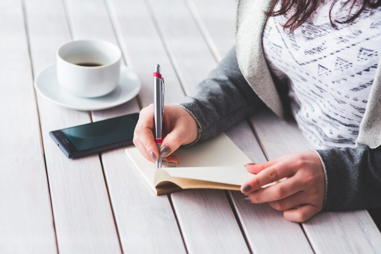 A woman writing in a journal next to a teacup and mobile phone.