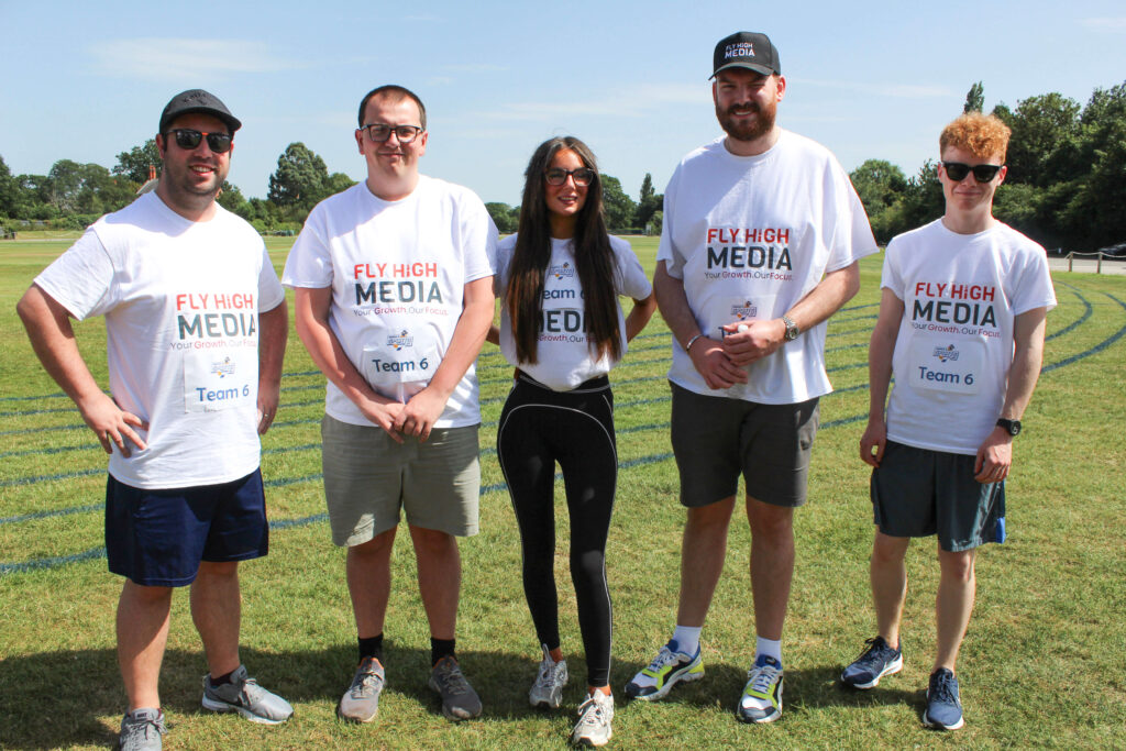 Fly High Media team members standing together on a sports field wearing branded T-shirts during a charity sports day event in sunny weather.