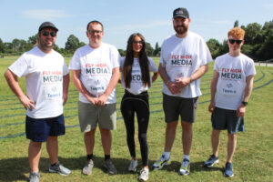Fly High Media team members standing together on a sports field wearing branded T-shirts during a charity sports day event in sunny weather.